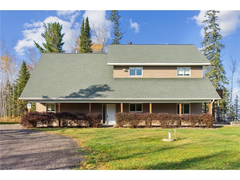 Two-story residential house with green roof, brick and tan siding, surrounded by evergreen trees and green lawn